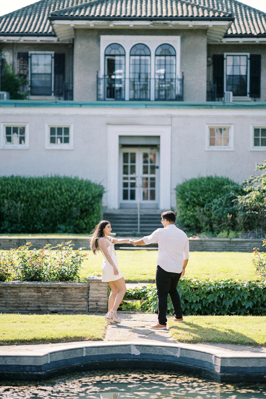 A couple enjoying a romantic moment in front of a large, elegant house with lush green garden surroundings.