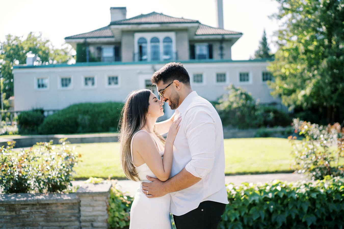 A couple embracing in front of a scenic house, surrounded by lush greenery on a sunny day.