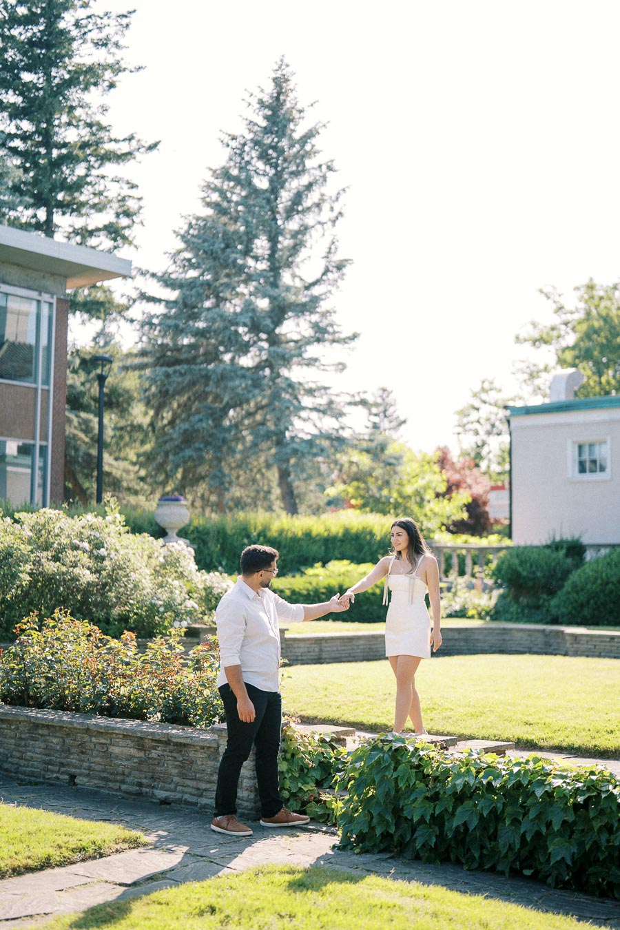 A couple enjoying a sunny day in a lush garden, with the man in a white shirt reaching out to the woman in a white dress, surrounded by greenery and a modern building in the background.