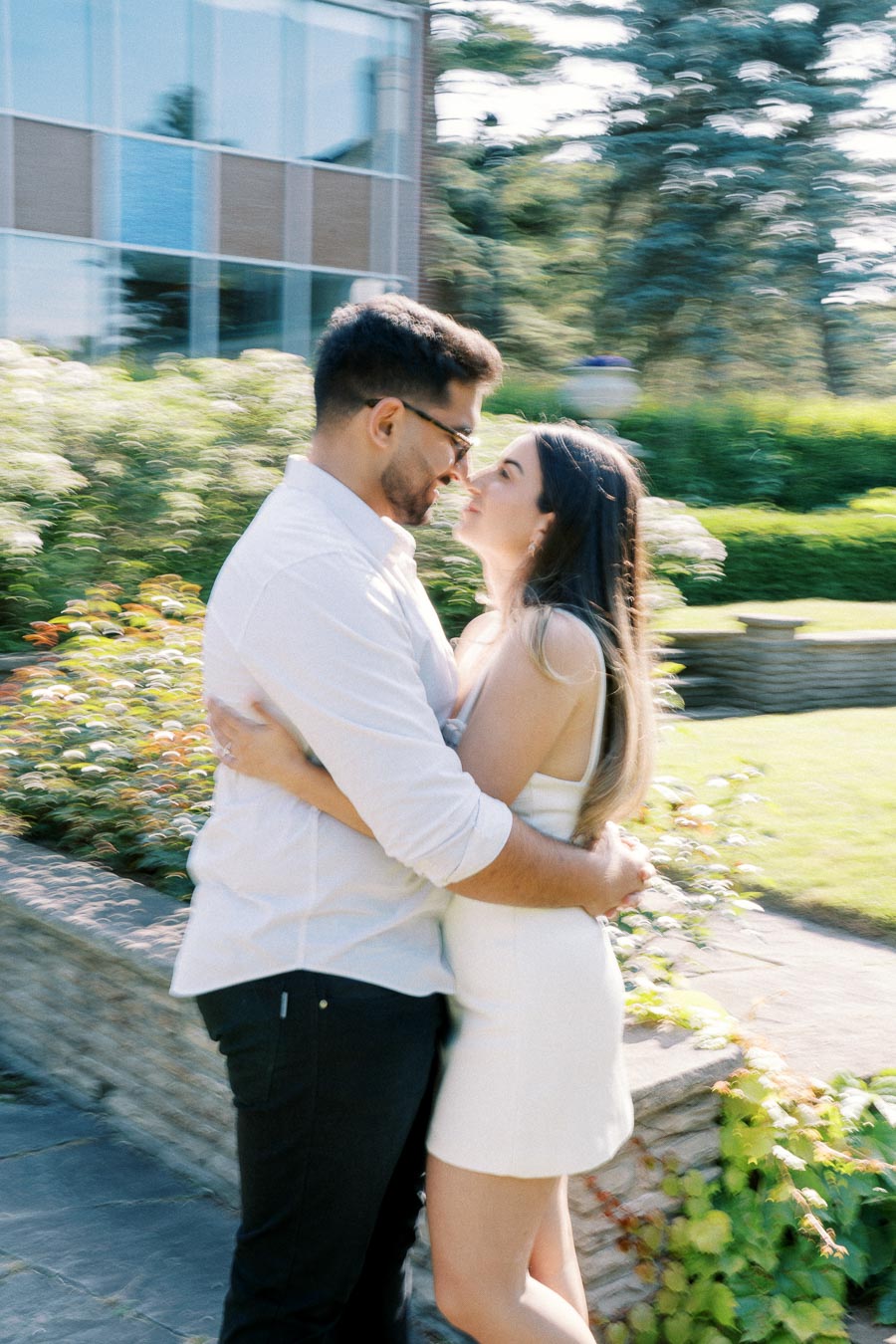 A couple embracing outdoors in a sunny garden setting, both smiling and looking into each other's eyes.