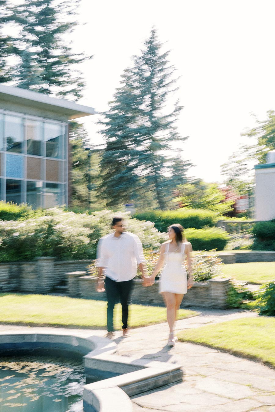 Blurred image of a couple holding hands while walking in a sunny garden near a modern building, surrounded by lush greenery and a small pond.