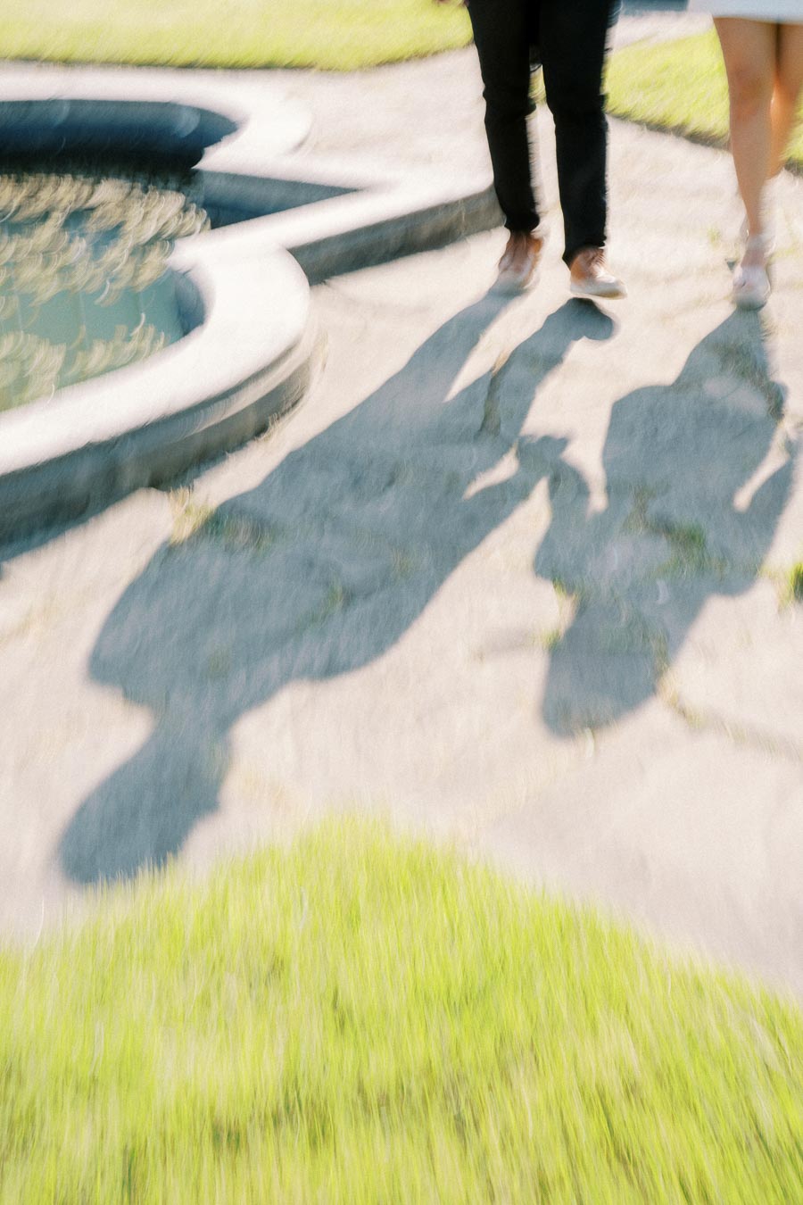 Blurred image of a couple walking hand in hand on a sunny day, casting long shadows on a stone pathway next to a curved water feature, surrounded by grass.