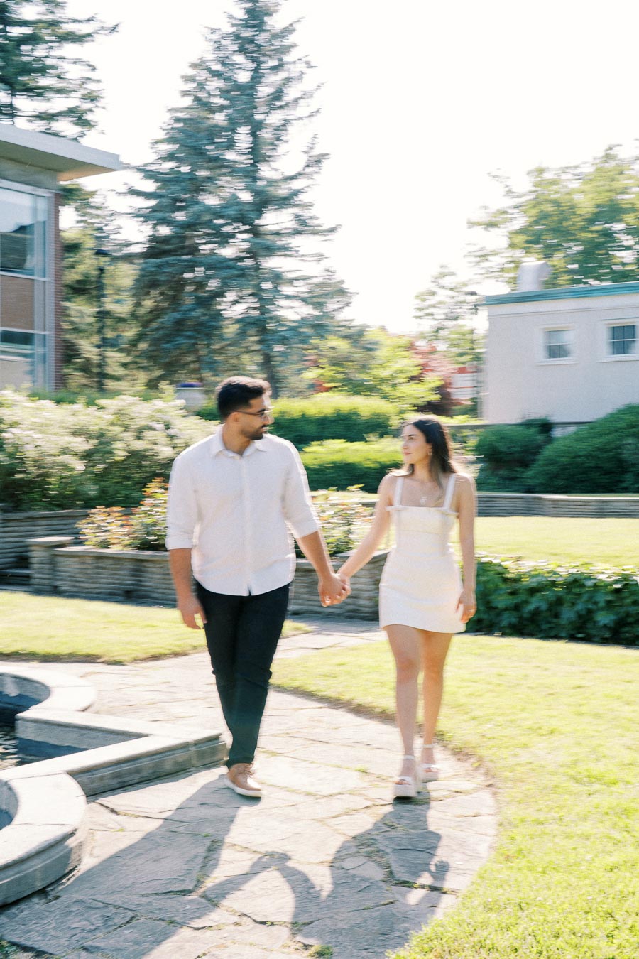 A couple holds hands while walking along a sunlit garden pathway, surrounded by greenery and trees, on a bright day.