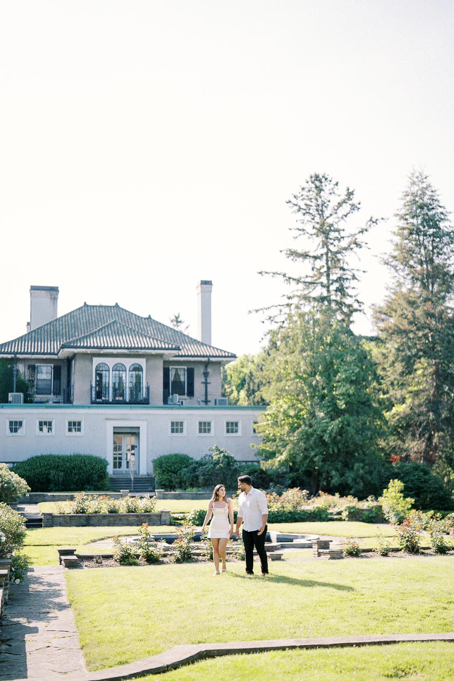 A couple walking in a lush garden in front of a grand two-story mansion with large windows and trees in the background, under a clear blue sky.