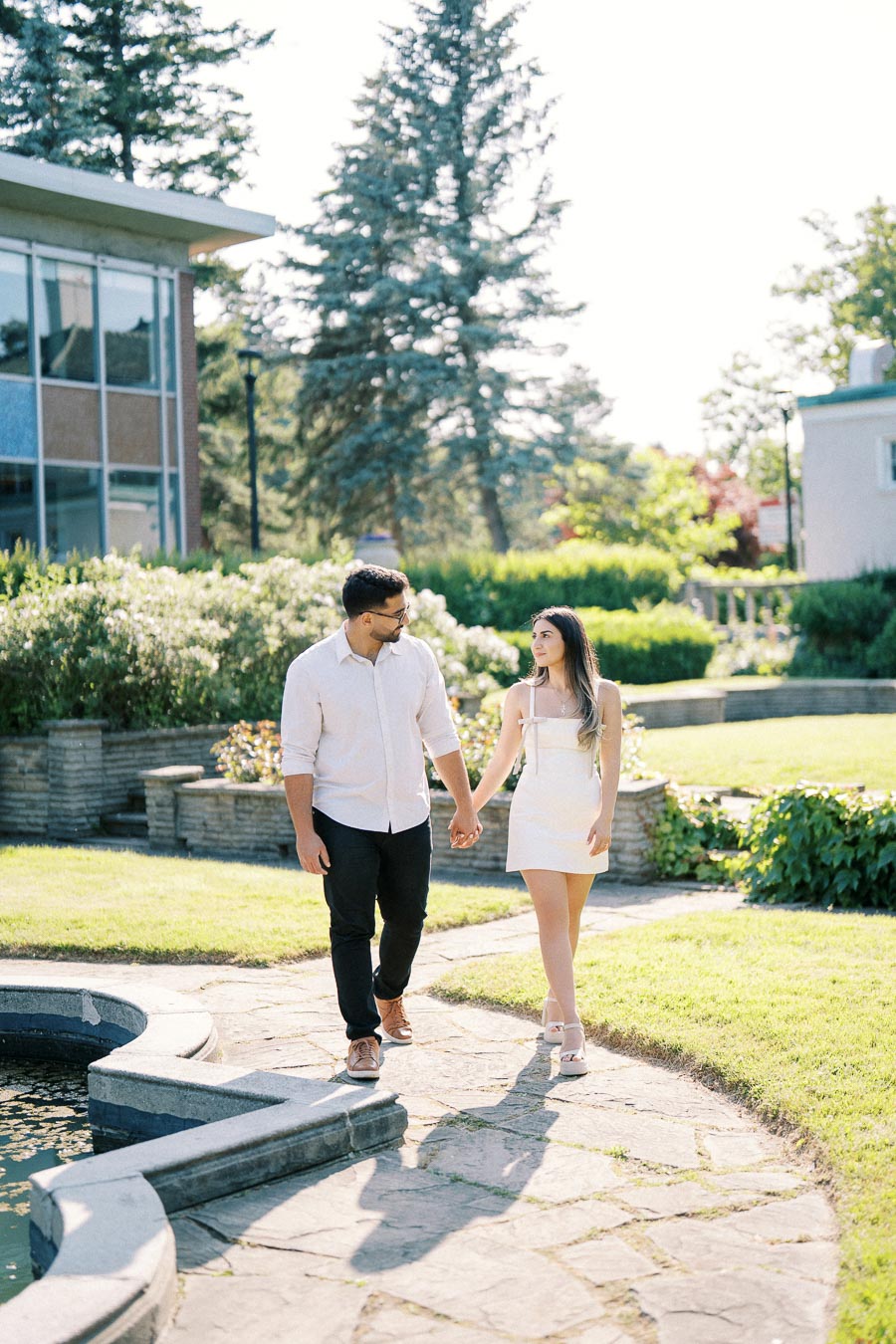 A couple holding hands and walking on a sunny pathway in a scenic garden setting, with lush greenery and modern buildings in the background.