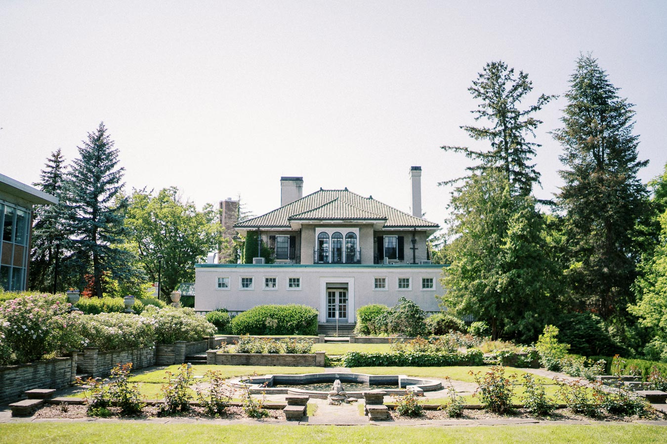 Elegant historical mansion surrounded by lush gardens and trees on a sunny day, featuring classic architectural design with a prominent entrance and fountain in the foreground.
