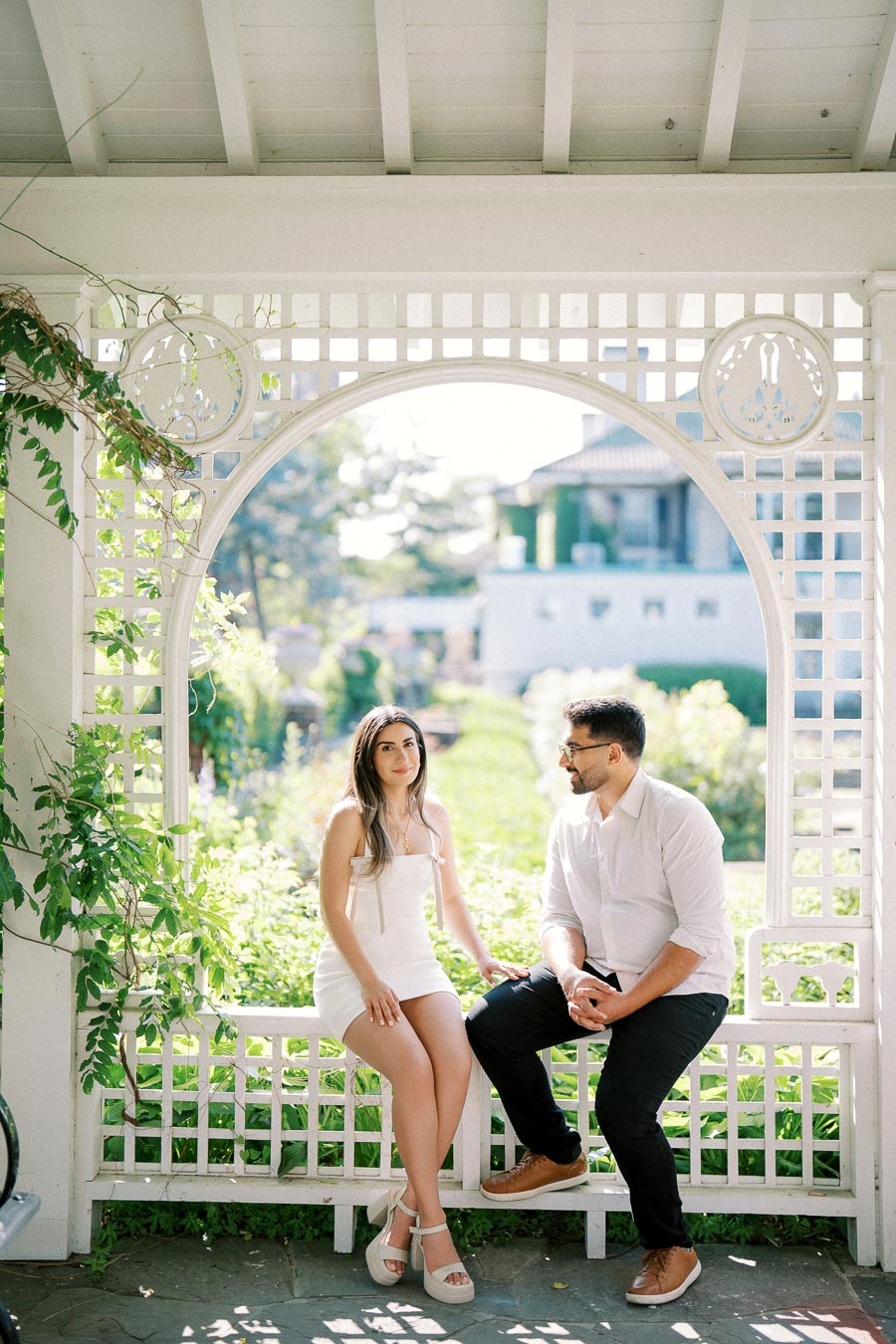 Couple sitting on a white wooden bench in a sunlit garden gazebo, surrounded by lush greenery, creating a serene and romantic atmosphere.
