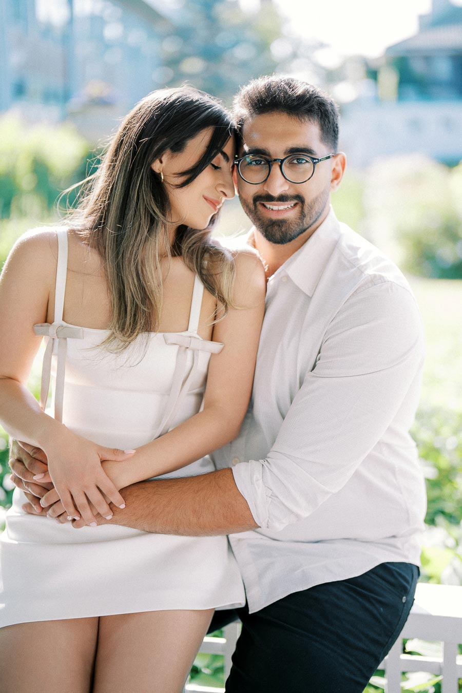 A happy couple embraces outside in a sunlit garden, both wearing casual summer attire and smiling warmly, capturing a romantic moment.