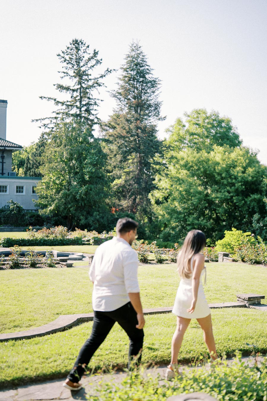 A couple walking through a lush garden with tall trees and a historic building in the background on a sunny day.