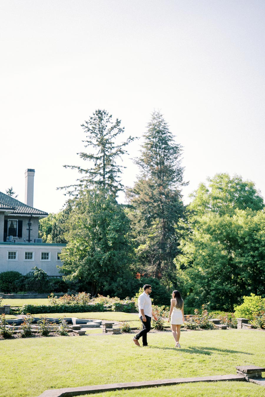A couple walking in a lush garden with a historic mansion and tall trees in the background on a sunny day.