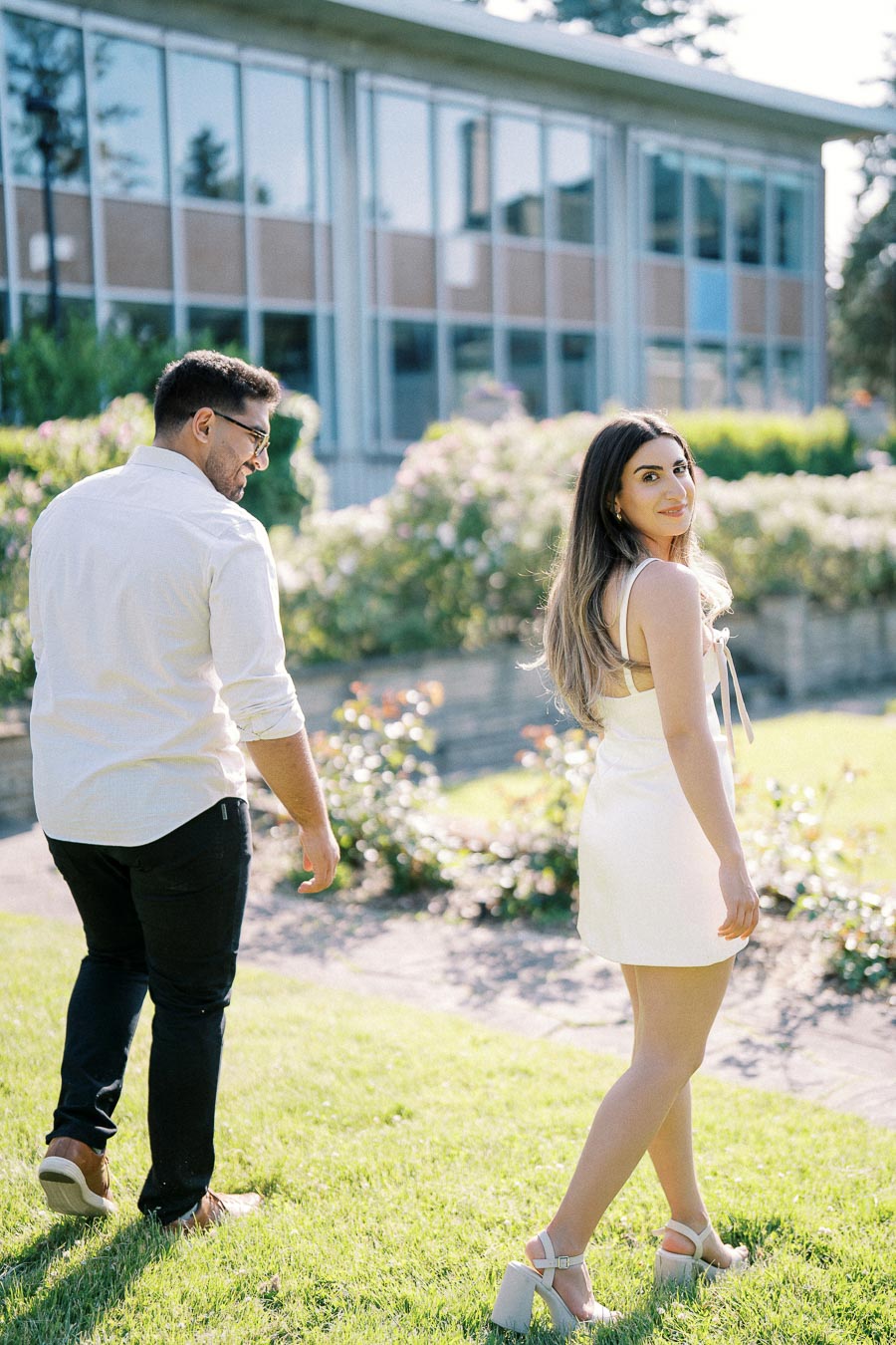 Couple walking on a grassy path in a garden with a modern glass building in the background, exuding a romantic and summery vibe.