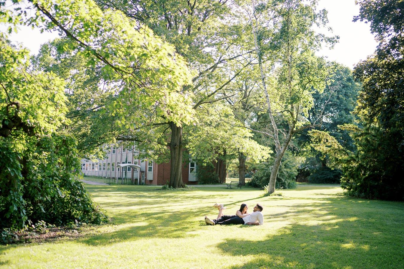 A couple relaxing on a sunlit green lawn surrounded by tall trees, with a red-brick building in the background on a bright summer day.