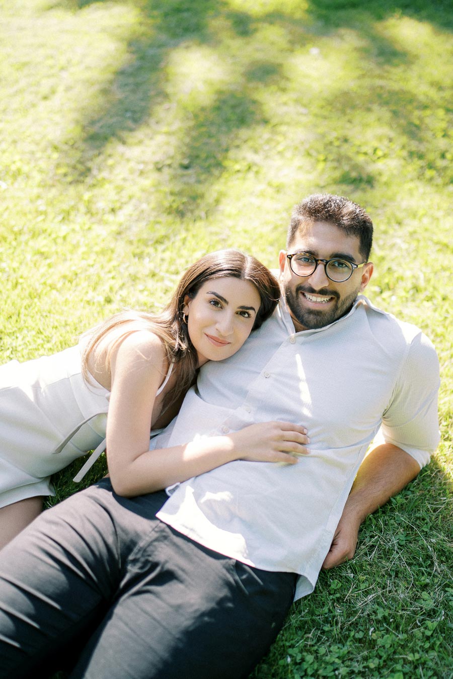 Couple lying on grass in park, woman in white dress resting her head on man's chest, both smiling at camera.