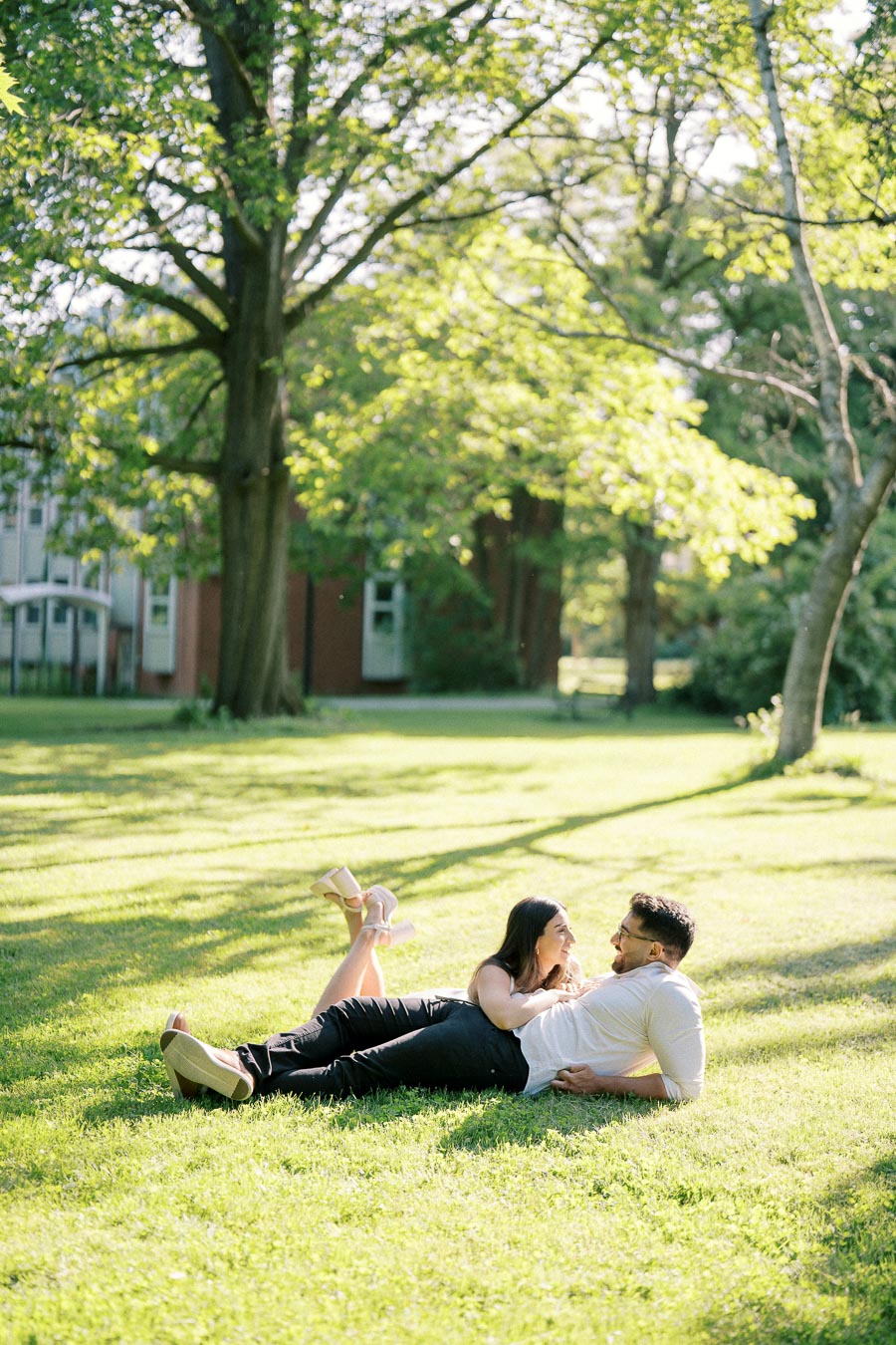A couple lying on a grassy lawn under a large tree, laughing and enjoying a sunny day in a park setting.