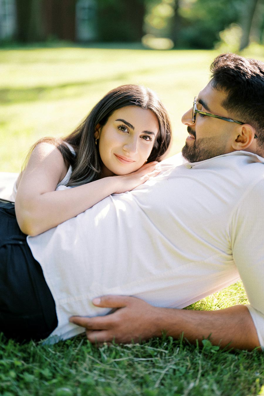A couple lying on grass in a sunny park, with the woman resting her head on the man's chest, both smiling contently.