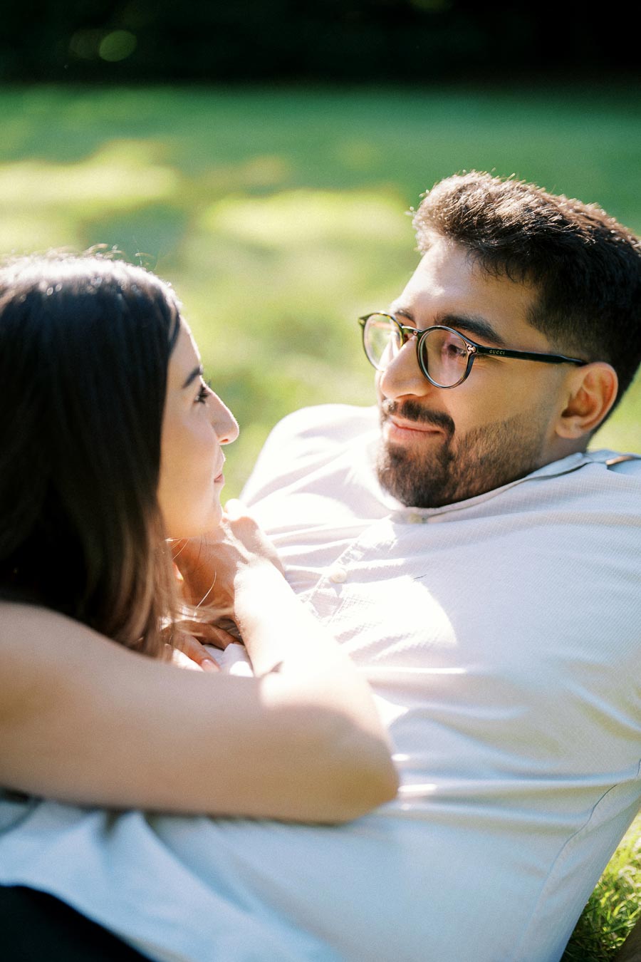 A couple lying on the grass, gazing into each other's eyes, surrounded by a lush green background on a sunny day.
