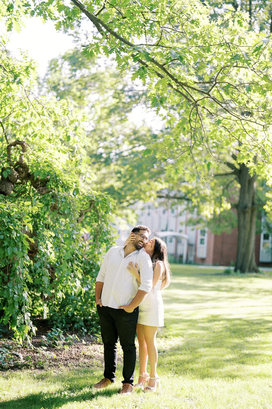 A couple embraces under lush green trees in a sunny park, with the woman gently kissing the man's cheek, expressing love and happiness in an outdoor setting.