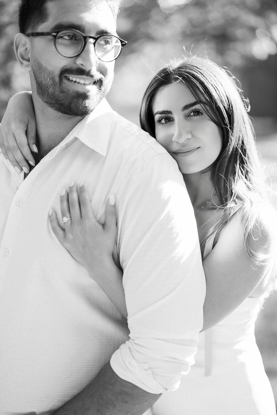 A couple embraces in a black and white photo, with a woman smiling gently while resting her hand on a man's shoulder, showcasing an engagement ring. The background is softly blurred with outdoor lighting.
