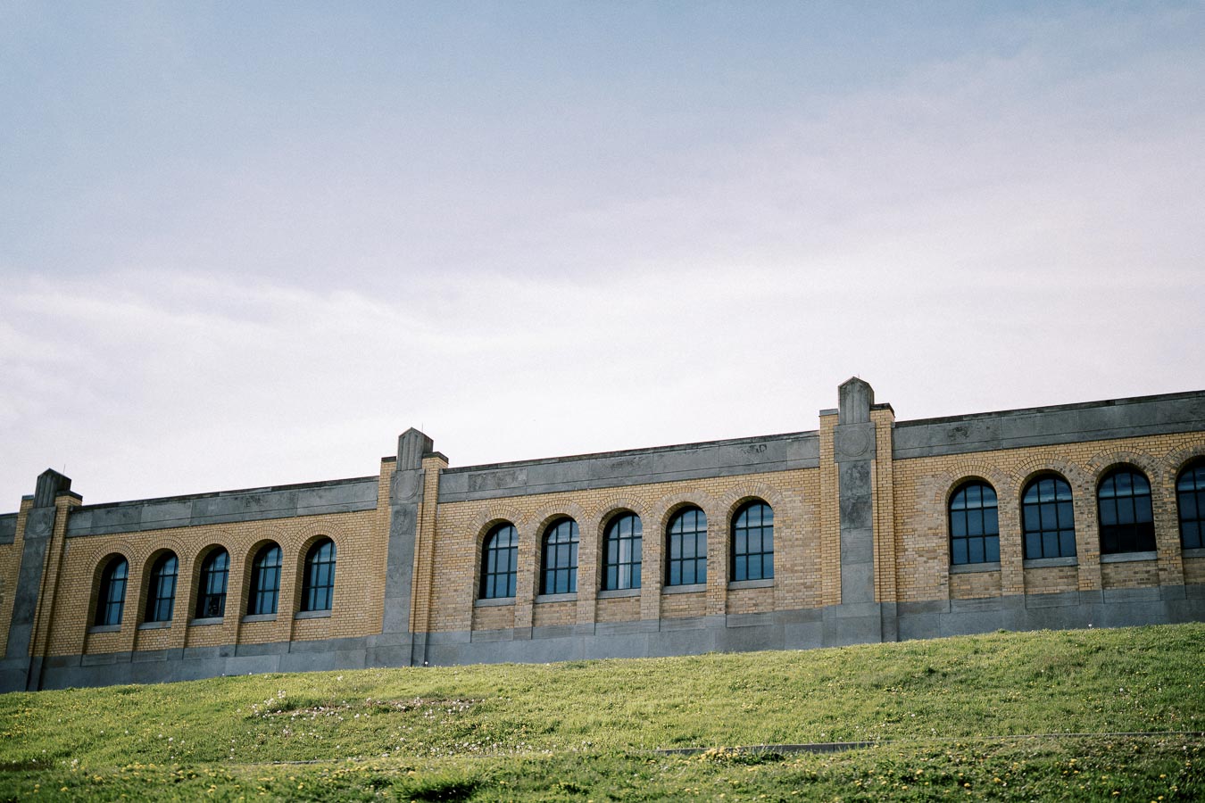Historic brick building with arched windows, partially obscured by a grassy hill under a clear sky.