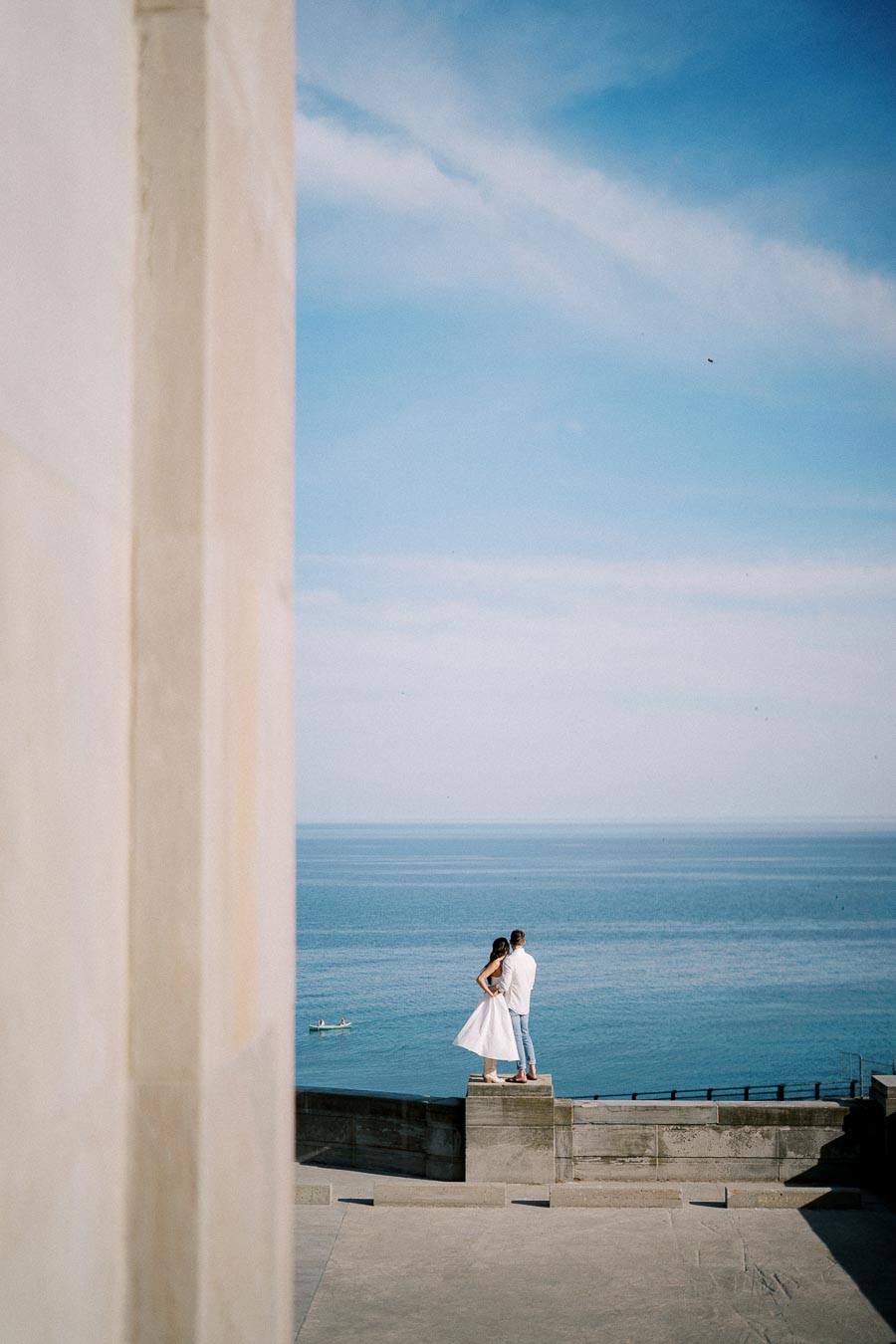 A couple stands on a concrete ledge overlooking a vast blue ocean under a clear sky, with a small boat visible in the distance, conveying a sense of romance and adventure.