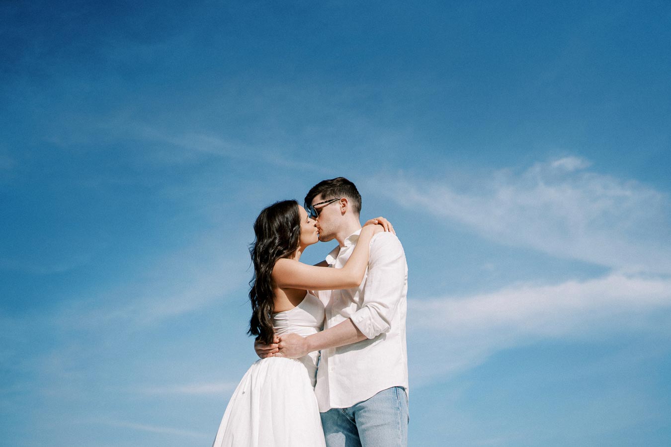 A couple in white attire kisses passionately under a clear blue sky, symbolizing love and romance.