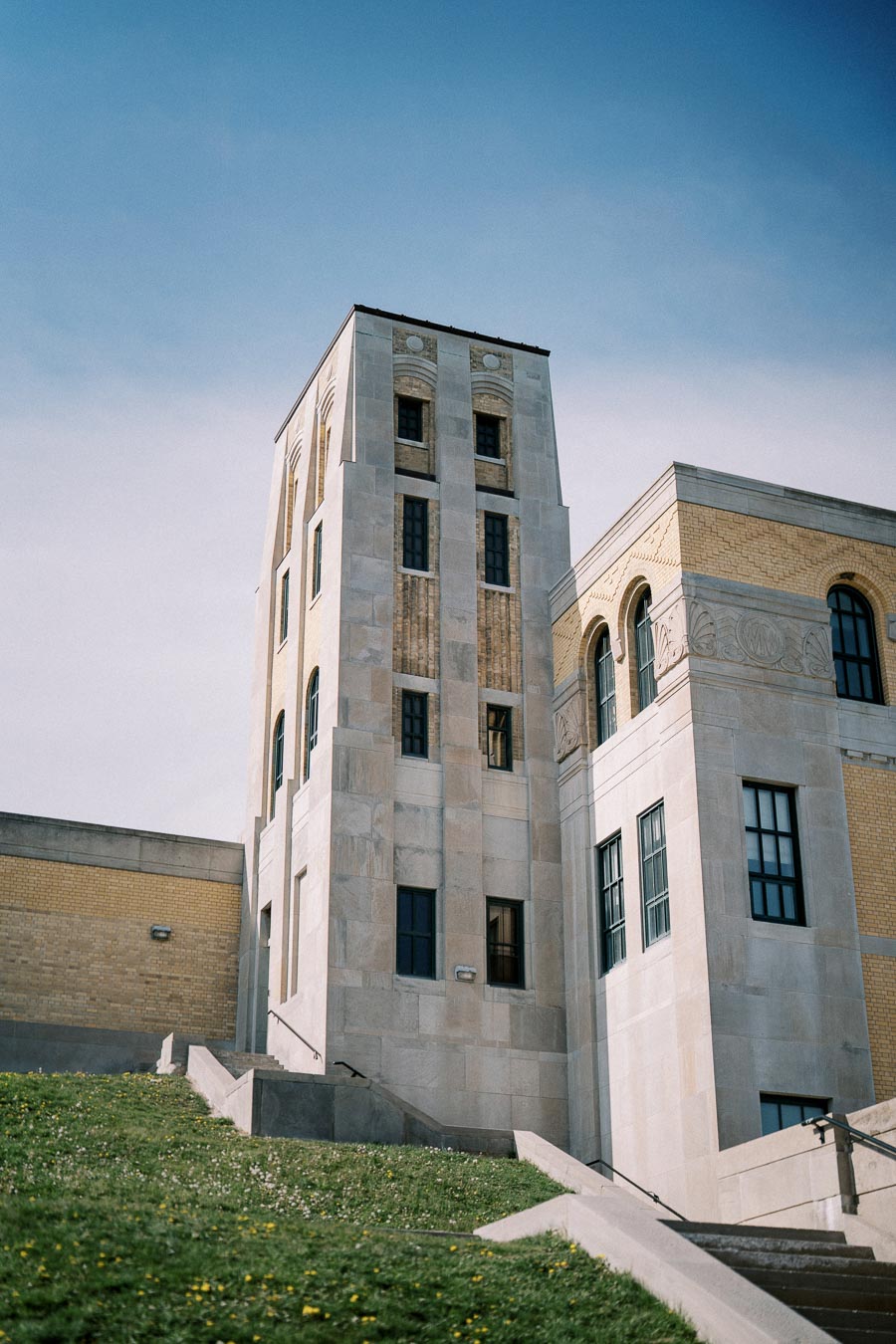 Historic stone and brick building with a tall tower under a clear blue sky, featuring classical architectural design elements and surrounded by green grass and dandelions.