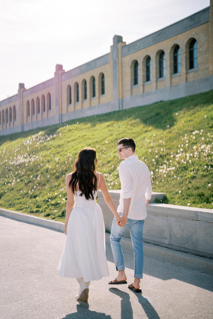 A couple walking hand in hand along a sunlit pathway, with a historical building and grassy hillside in the background, conveying romance and tranquility.