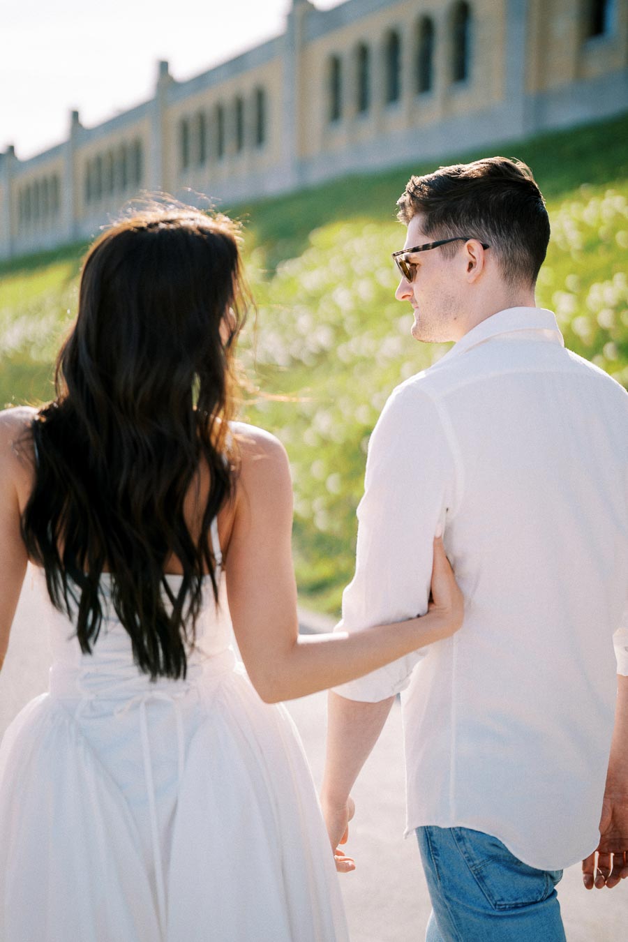 Couple walking outdoors on a sunny day, the woman in a white dress and the man in a white shirt, surrounded by greenery and architecture.
