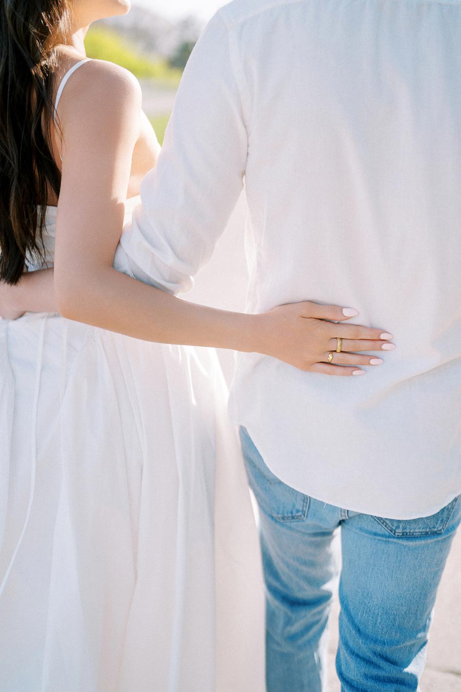 A couple embracing while walking, with the woman in a white dress and the man in a white shirt and blue jeans, captured from behind on a sunny day.