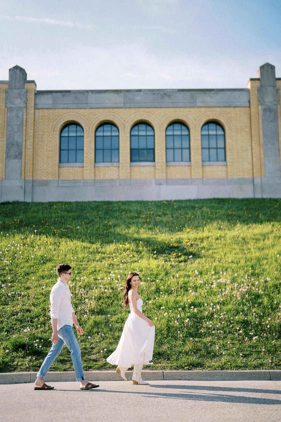 A couple walks hand in hand in front of a historic building with arched windows, set against a lush green hill, under a clear blue sky.