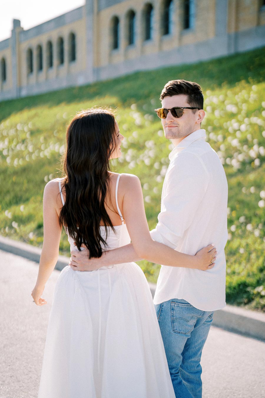A young couple smiling at each other while walking outside on a sunny day, with a backdrop of green grass and a building.