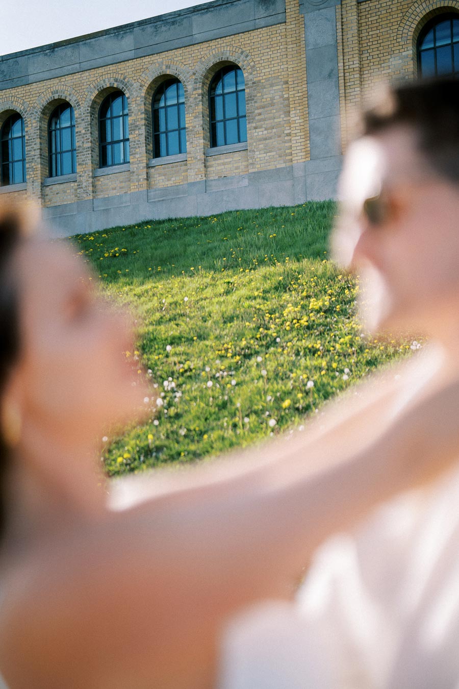 Blurred image of two people outdoors with a historic brick building in the background and a grassy field with flowers in the foreground.