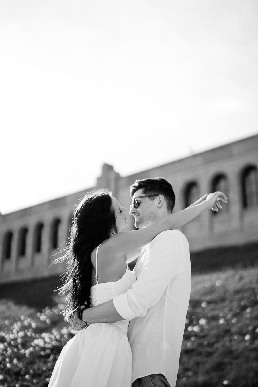 Black and white photo of a couple embracing outdoors with an architectural background. The woman is wearing a dress, and the man is wearing a shirt and sunglasses, conveying a romantic, timeless moment.