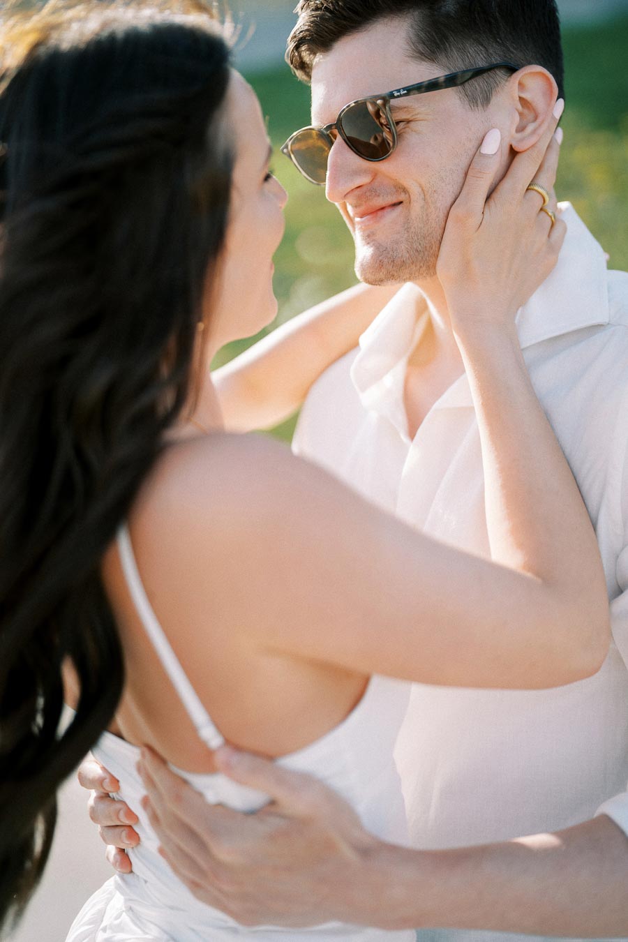 A couple embracing and smiling at each other outdoors on a sunny day, with the woman gently holding the man's face.