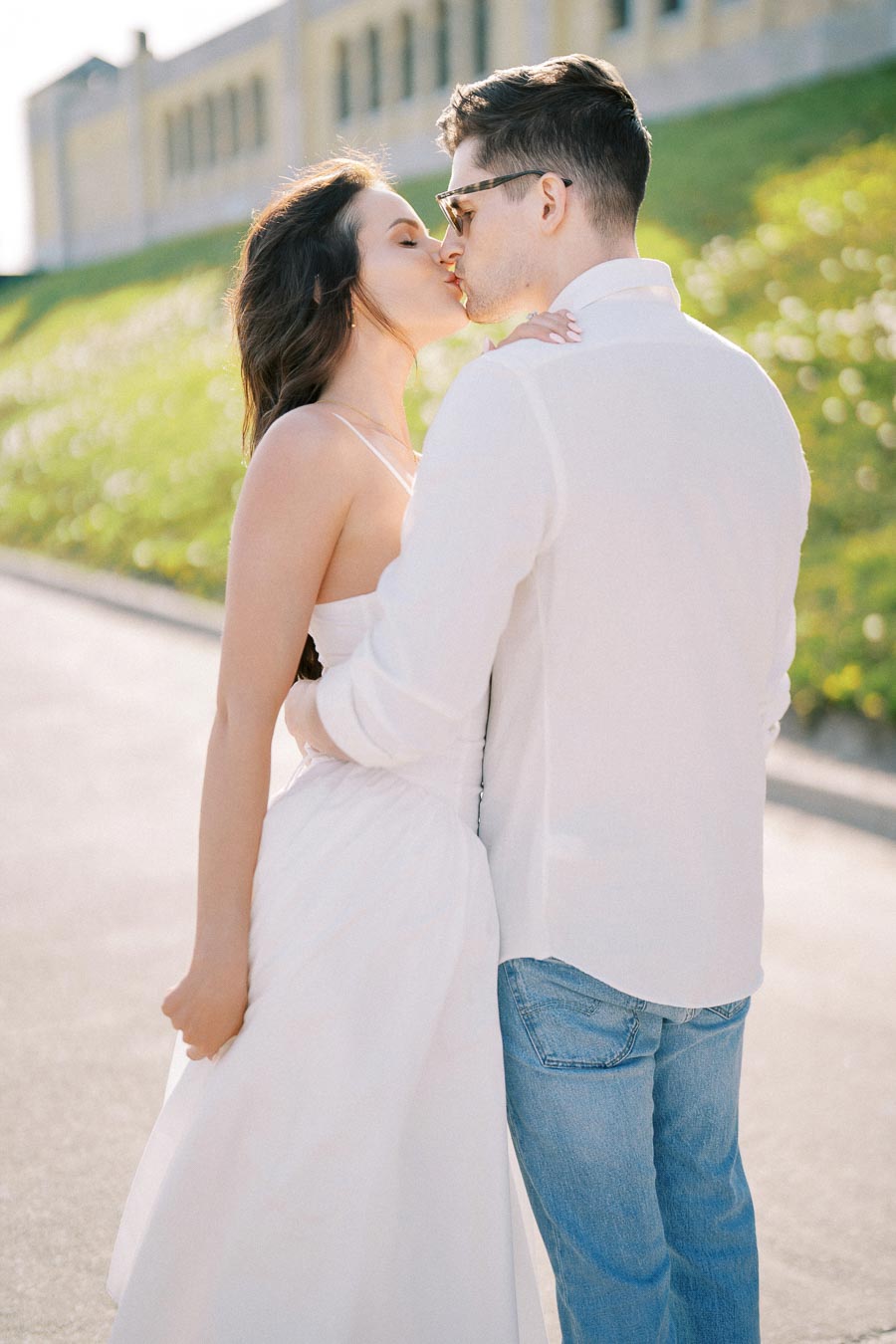 A couple shares a romantic kiss outdoors on a sunny day, with the woman in a white dress and the man in casual attire.