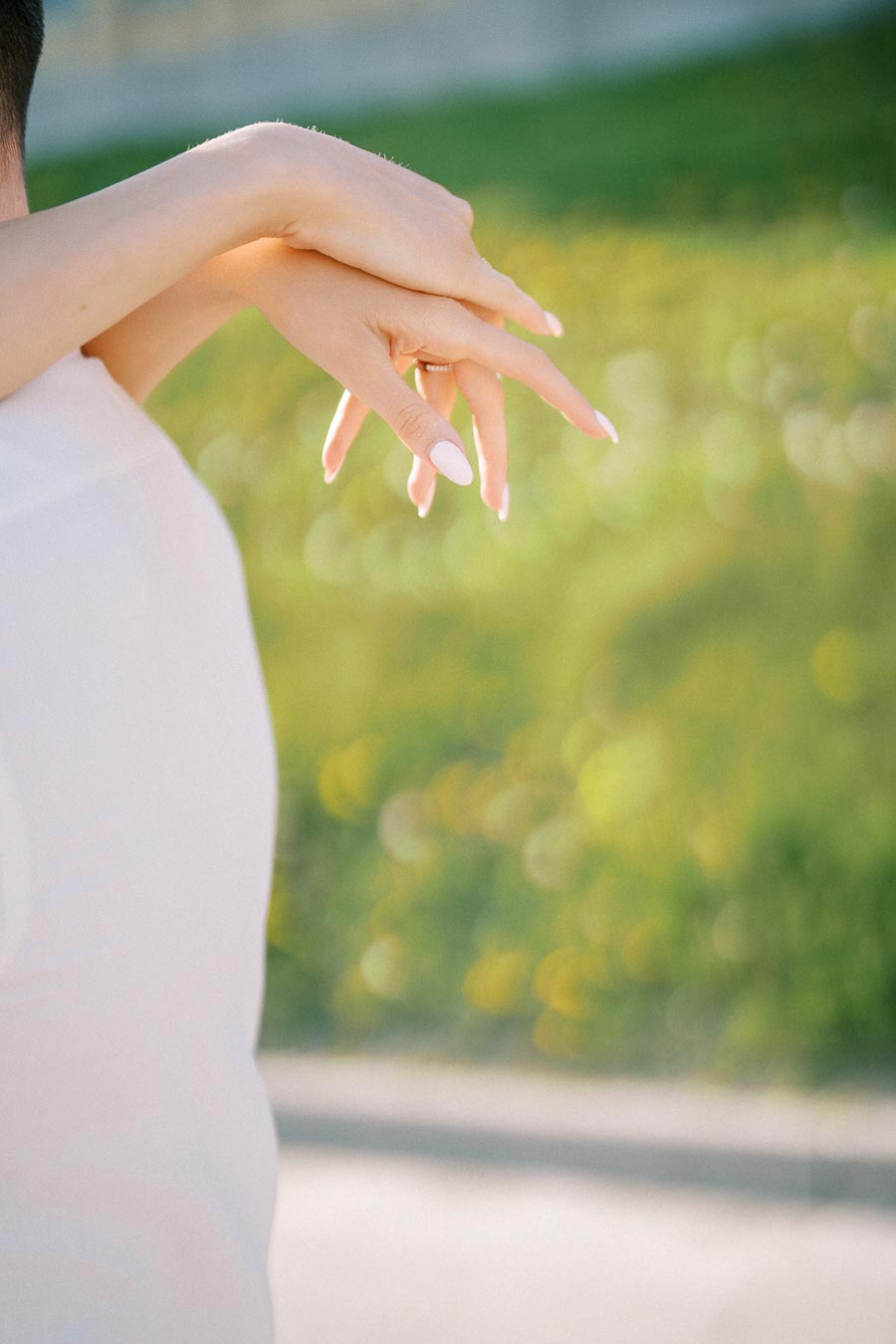 A close-up of a woman's hands with neatly manicured nails, resting on a man's shoulder, in an outdoor setting with a blurred green background.
