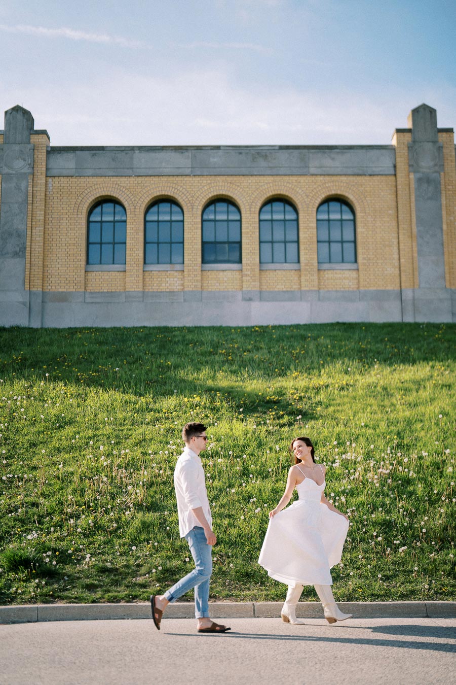 A couple walking joyfully in front of a historic yellow brick building with arched windows, surrounded by a lush green lawn and a clear blue sky.