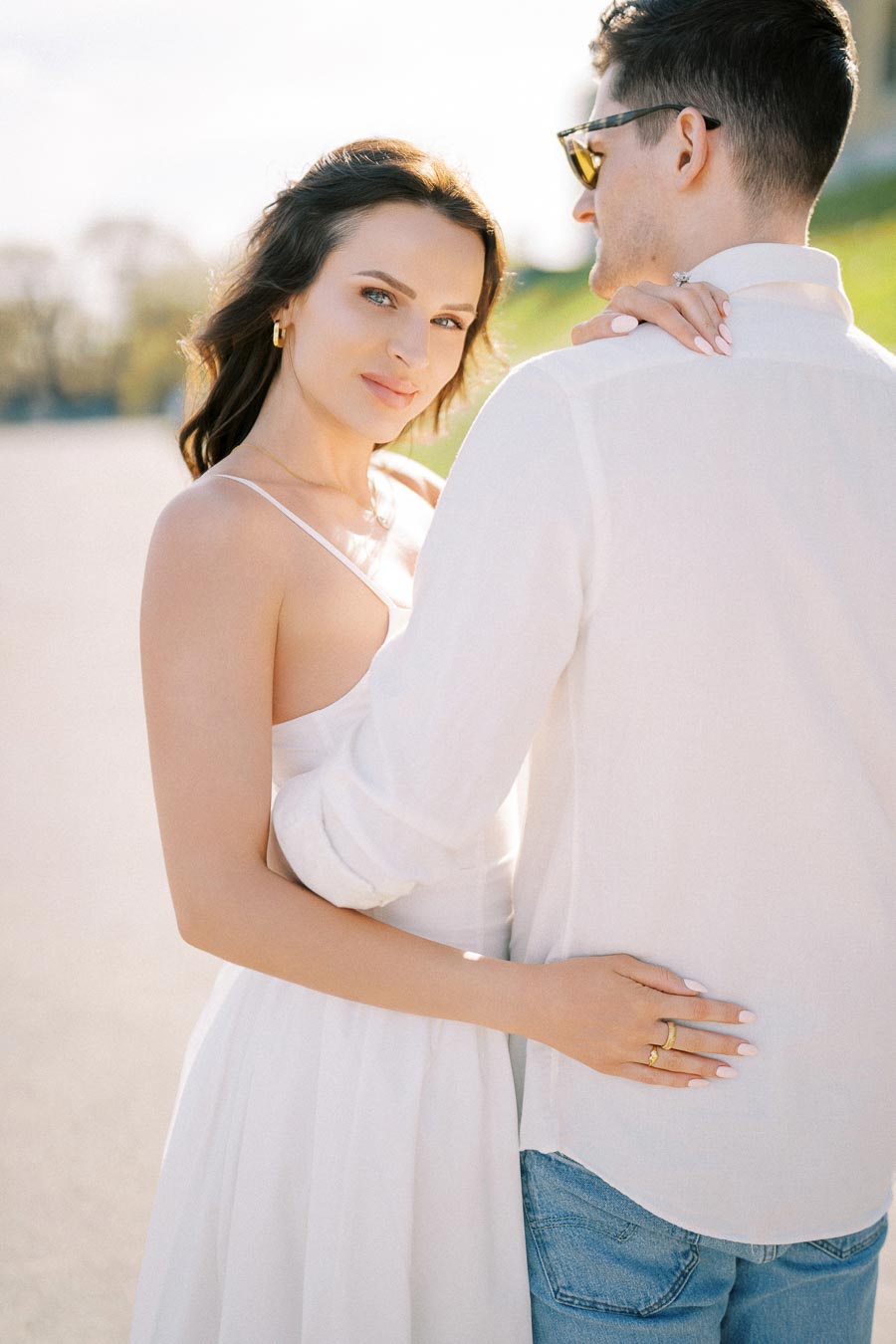 A woman in a white dress embraces a man wearing a white shirt and jeans, standing close together outdoors with a blurred background, capturing a romantic and sunny moment.