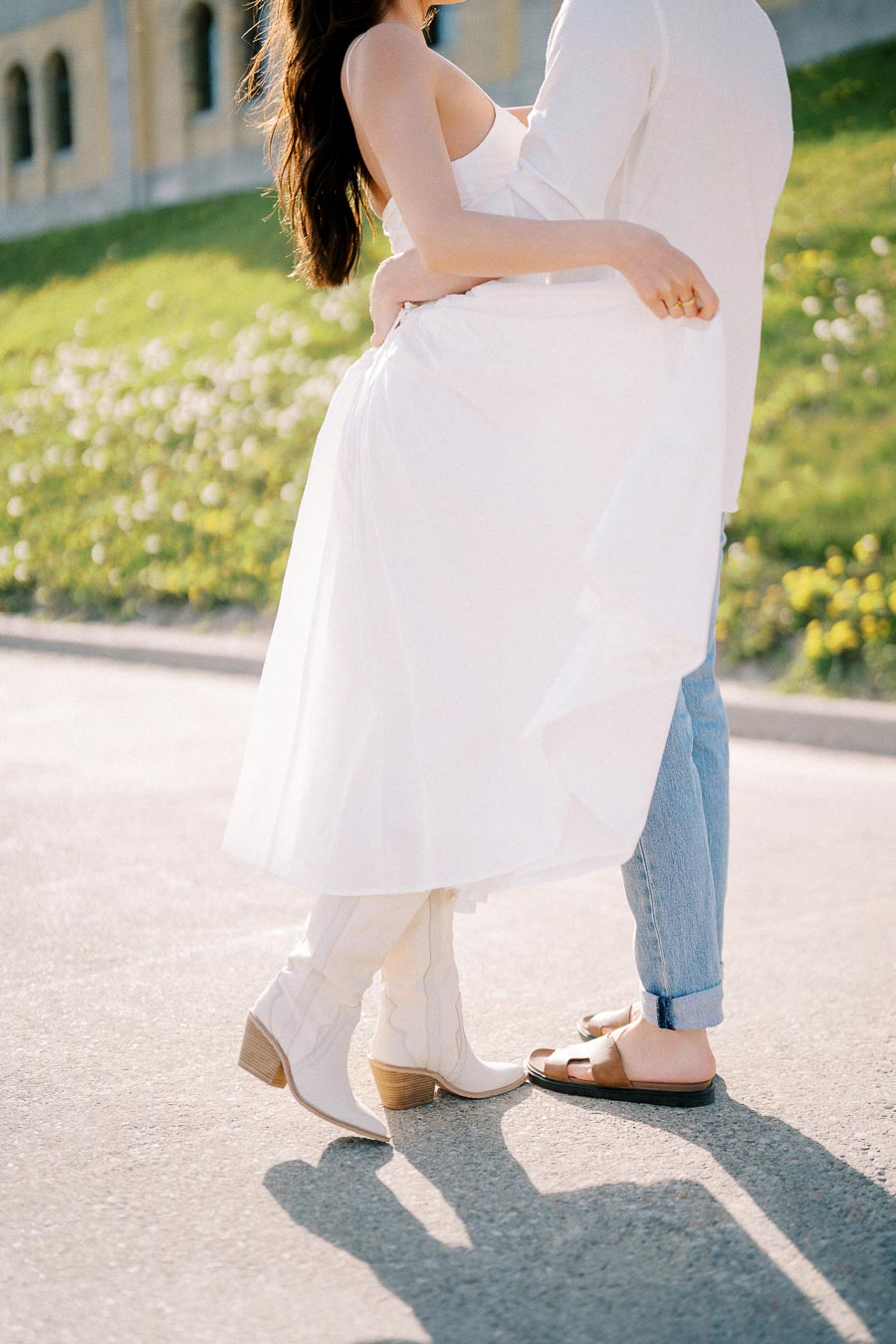 A couple embracing outdoors, with the woman wearing a flowing white dress and boots, and the man in casual jeans and sandals, standing on a sunlit pathway with a grassy background.