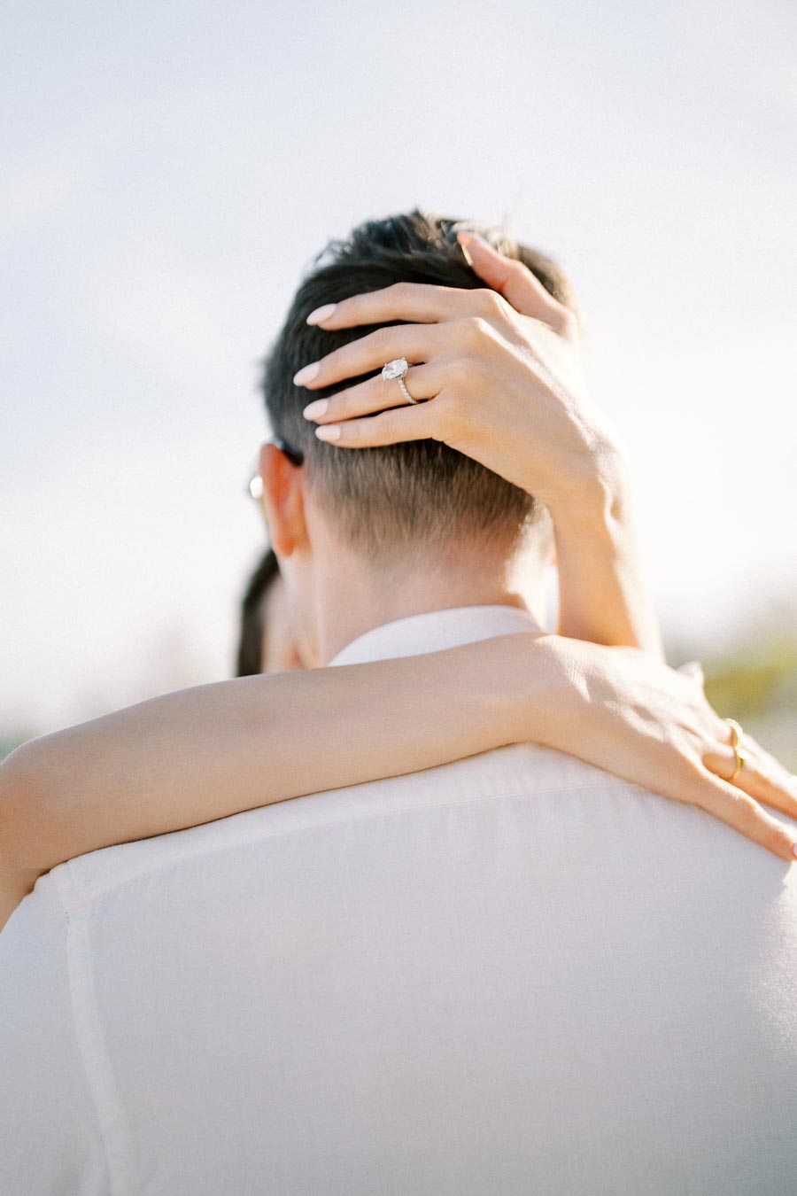 A couple embraces against a soft, light-filled background. Focus is on a woman's hand with an engagement ring resting on the man's neck. The sunlight highlights the elegant ring and the intimacy of the moment.