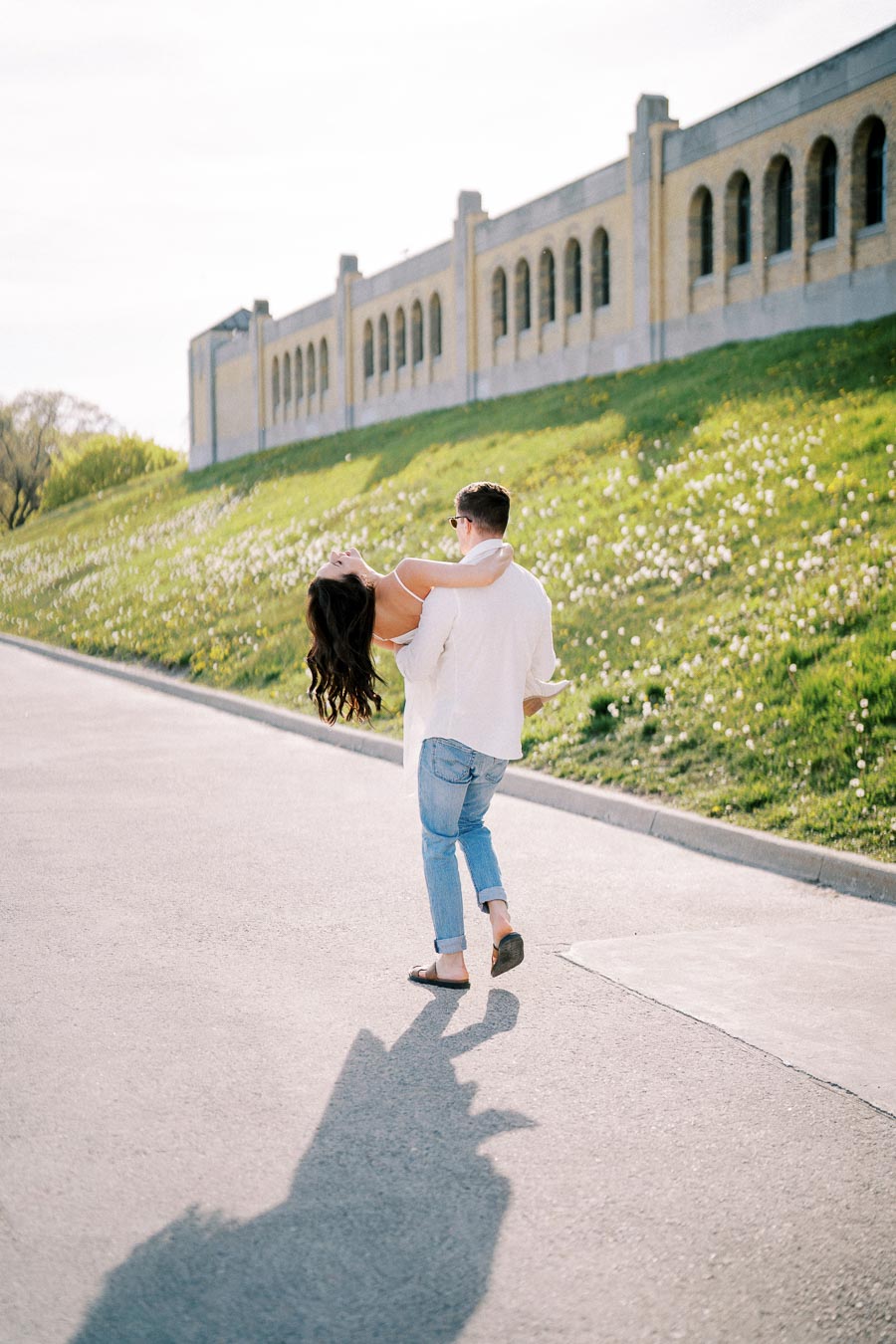 A man in casual summer attire carries a woman in a white dress along a sunlit path, with a historic building and green hillside in the background.