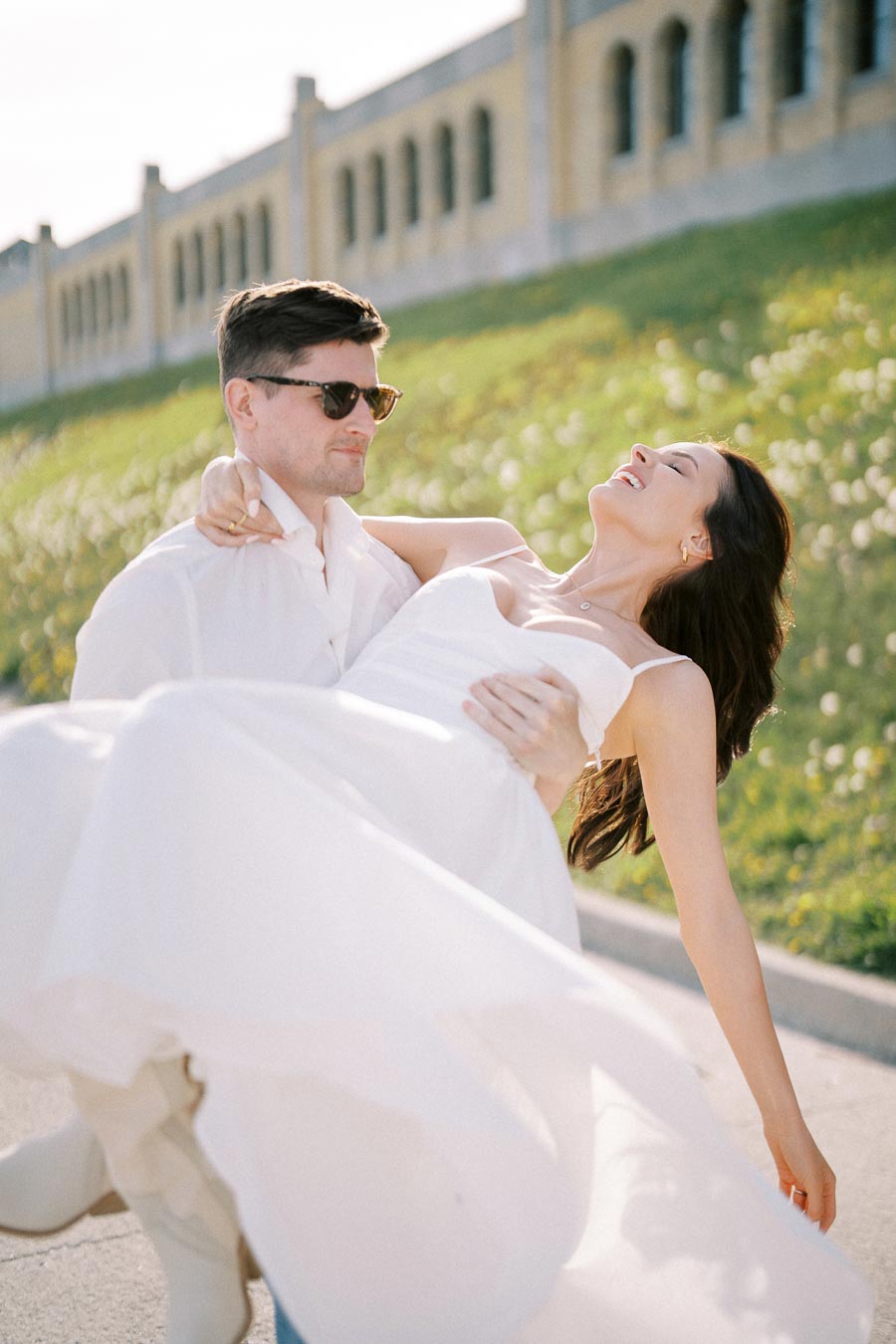 A joyful couple enjoys a sunny day, with a man carrying a smiling woman in a white dress near a historic building with a lush, green landscape in the background.