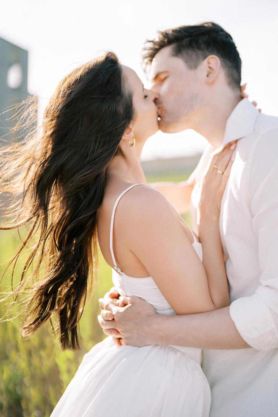 A romantic couple embracing and sharing a kiss in a sunlit outdoor setting, with the woman's hair flowing in the breeze and soft focus greenery in the background.