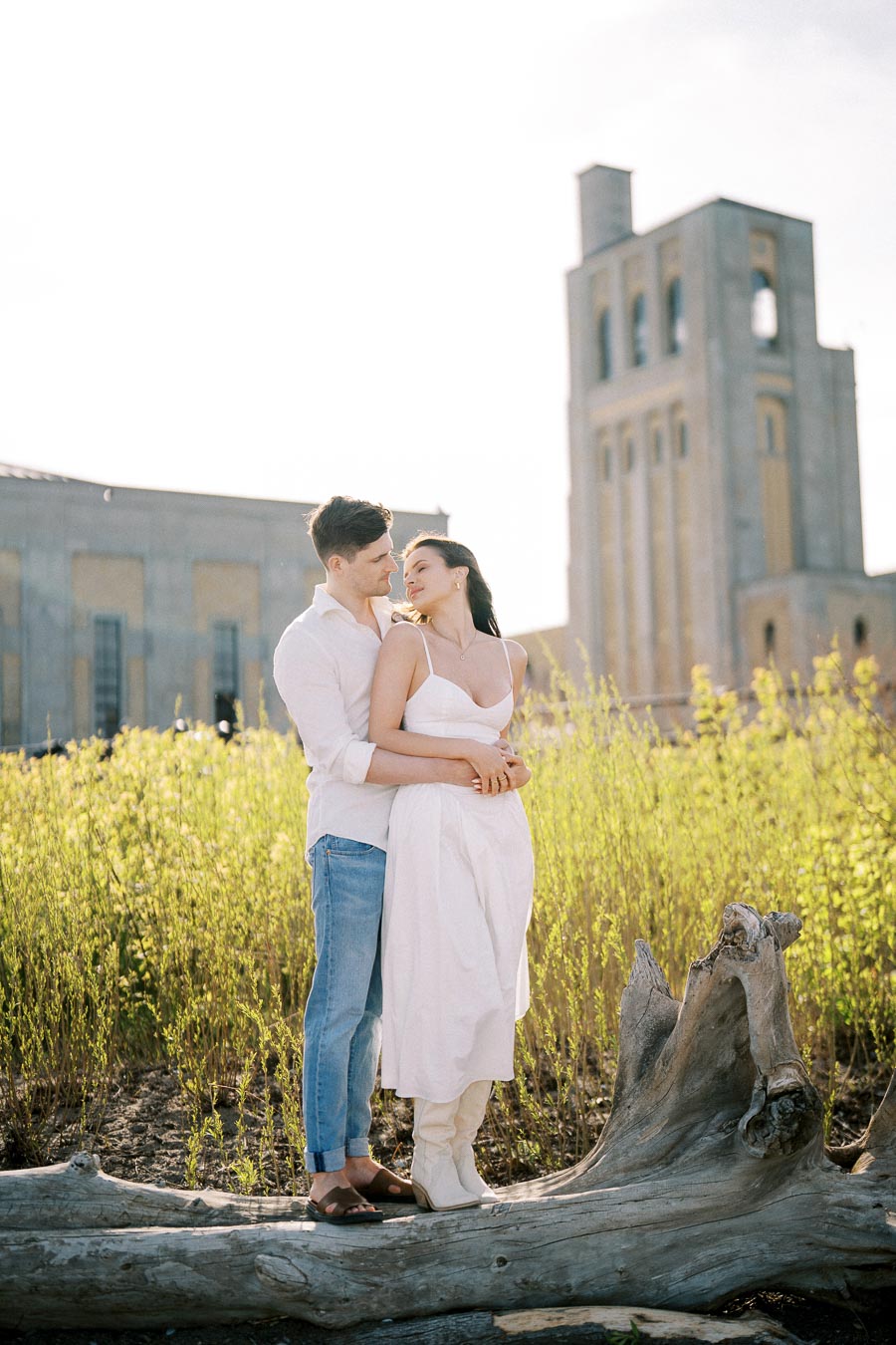 A couple embracing in a field with a tall, historical building in the background, captured under soft daylight, conveying romance and serenity.
