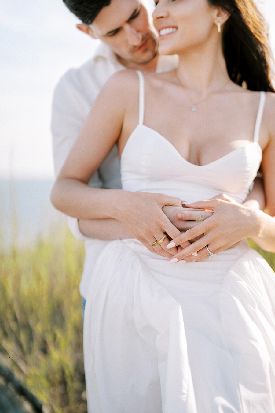 A couple in an intimate embrace outdoors, with the woman wearing a white dress and the man gently holding her from behind, highlighting romance and connection.