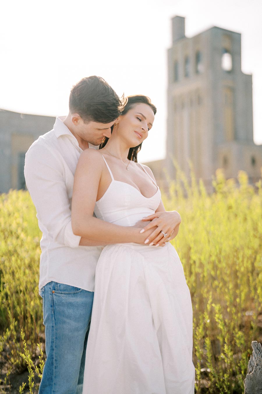 A couple embracing in a sunlit field with an old stone building in the background, wearing casual white outfits.