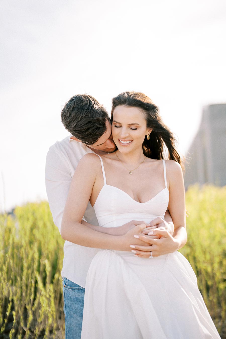 Romantic couple embracing in a sunlit field, both wearing white outfits, enjoying an intimate moment.