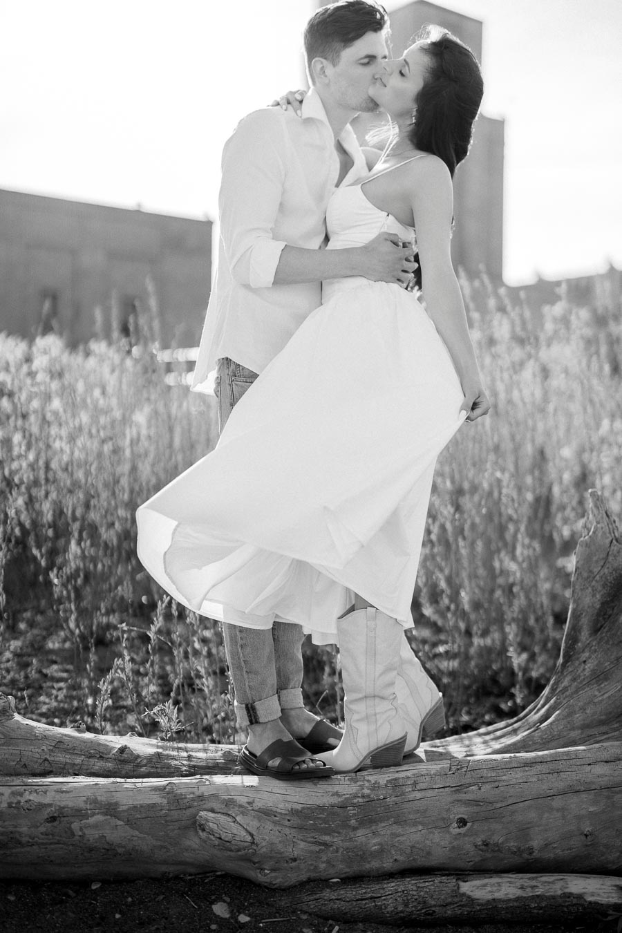 A black and white photo of a romantic couple sharing a kiss outdoors, standing on a log in a field. The woman wears a flowing white dress and cowboy boots, while the man is in casual attire. Sunlight highlights the natural setting, creating a serene atmosphere.