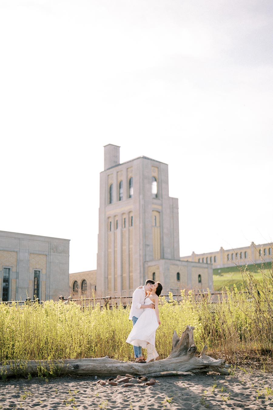 A couple embracing in front of a large, historic building surrounded by greenery, with a clear sky above.