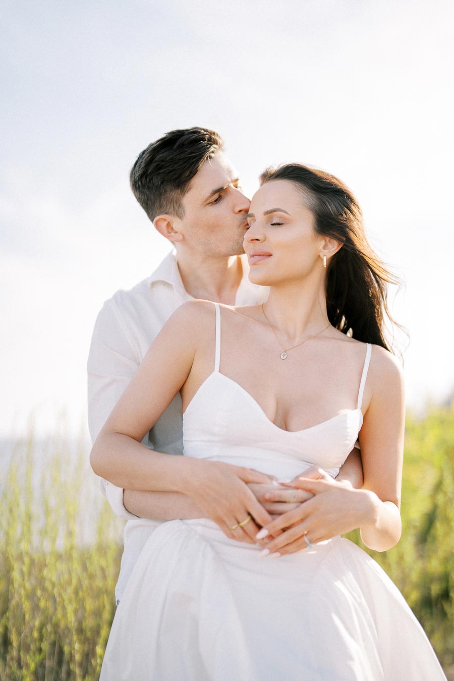 A couple embracing affectionately, with the man kissing the woman's temple in a serene, sunlit outdoor setting, both wearing white attire.